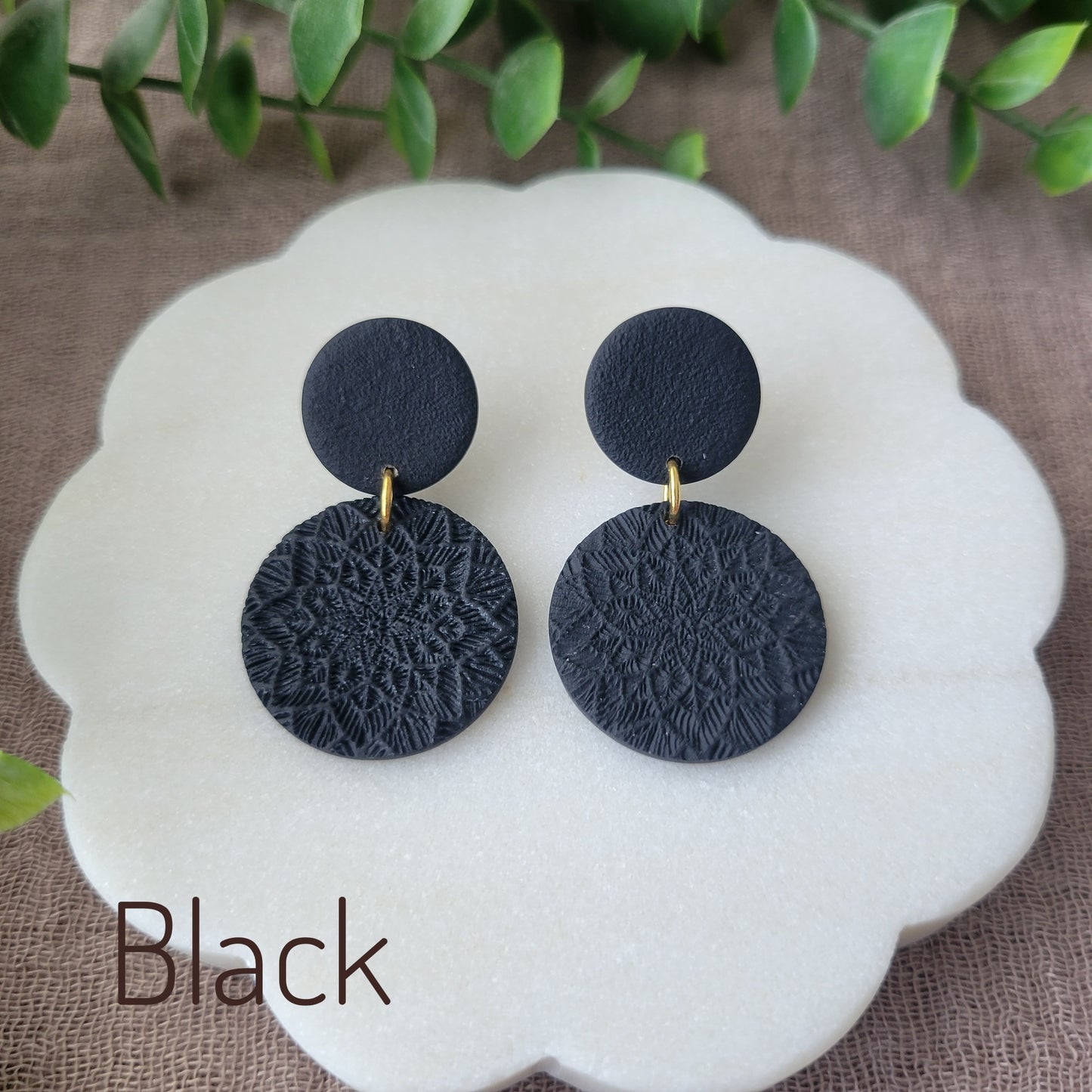 Top view of a pair of black mandala textured earrings displayed on a white round scalloped tray.