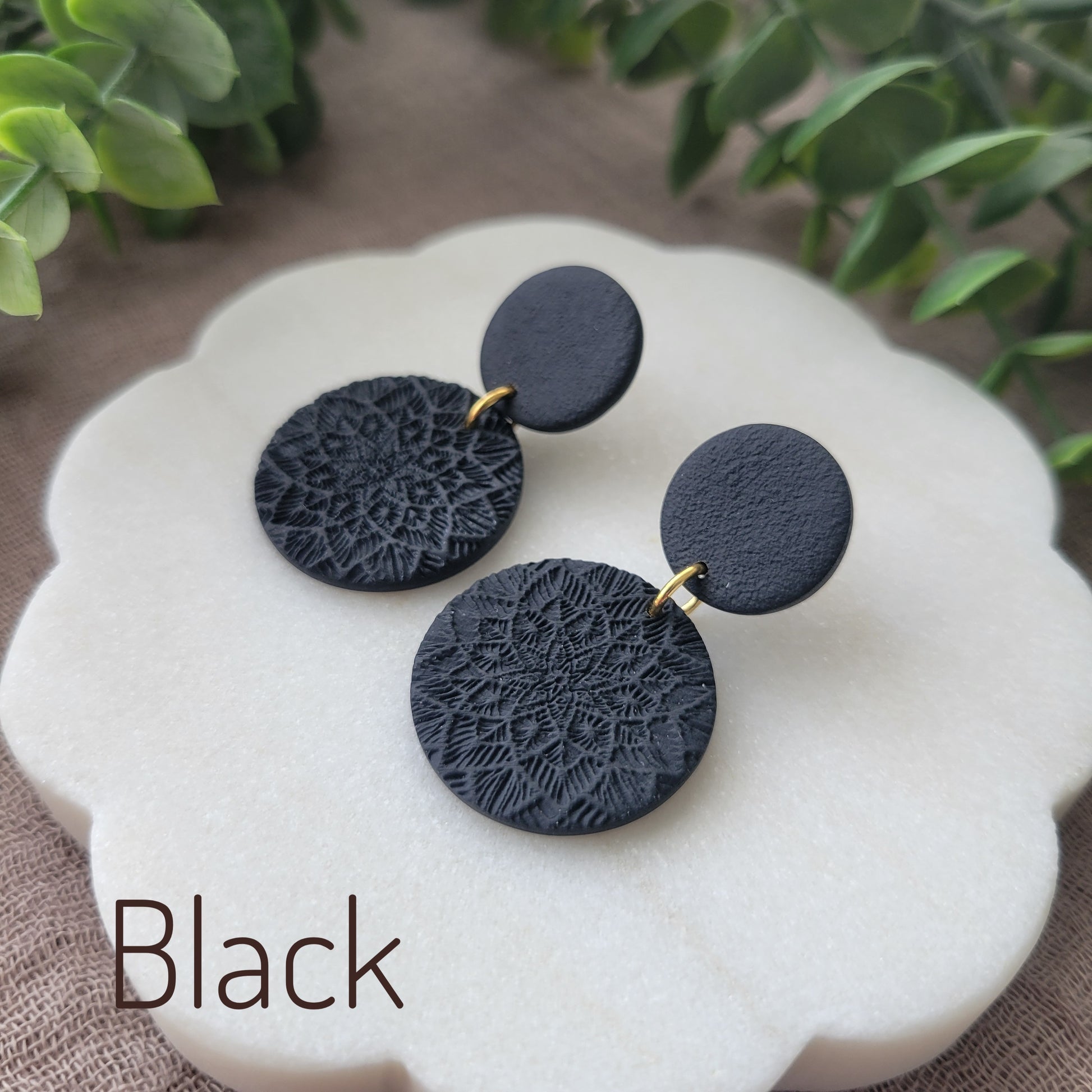 Side view of a pair of black mandala textured earrings displayed on a white round scalloped tray.