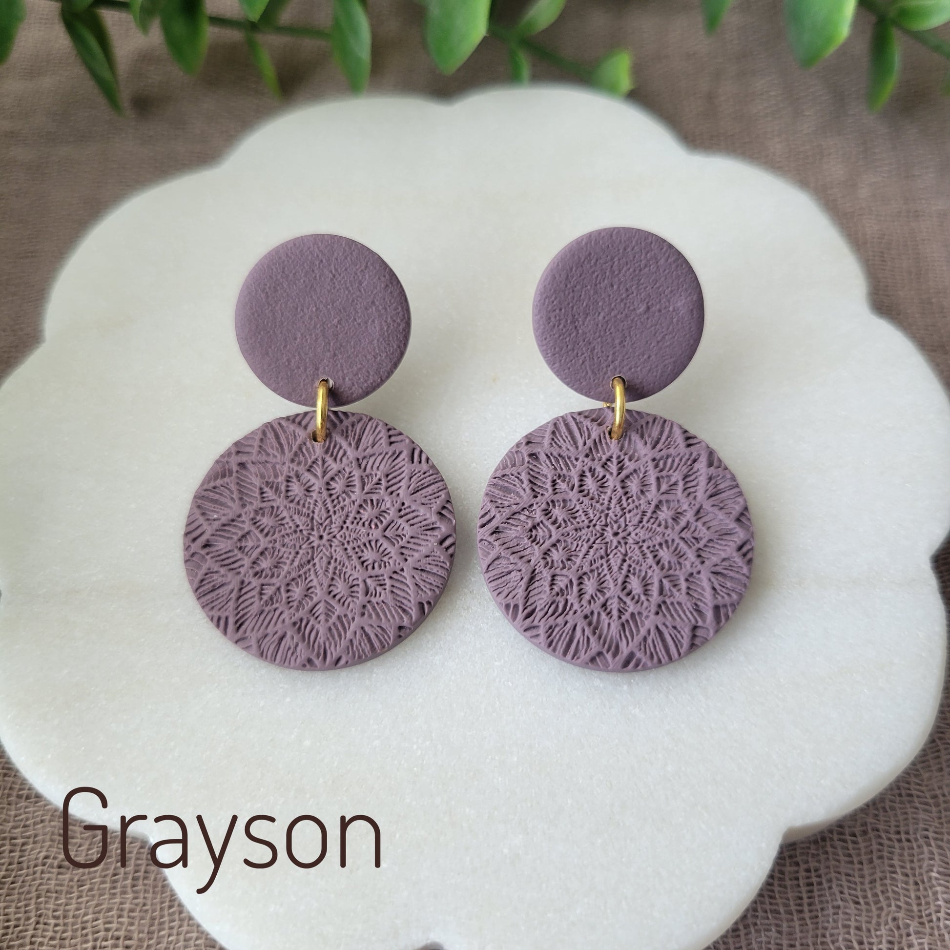 Top view of a pair of purple mandala textured earrings displayed on a white round scalloped tray.