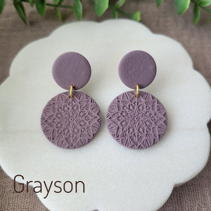 Top view of a pair of purple mandala textured earrings displayed on a white round scalloped tray.