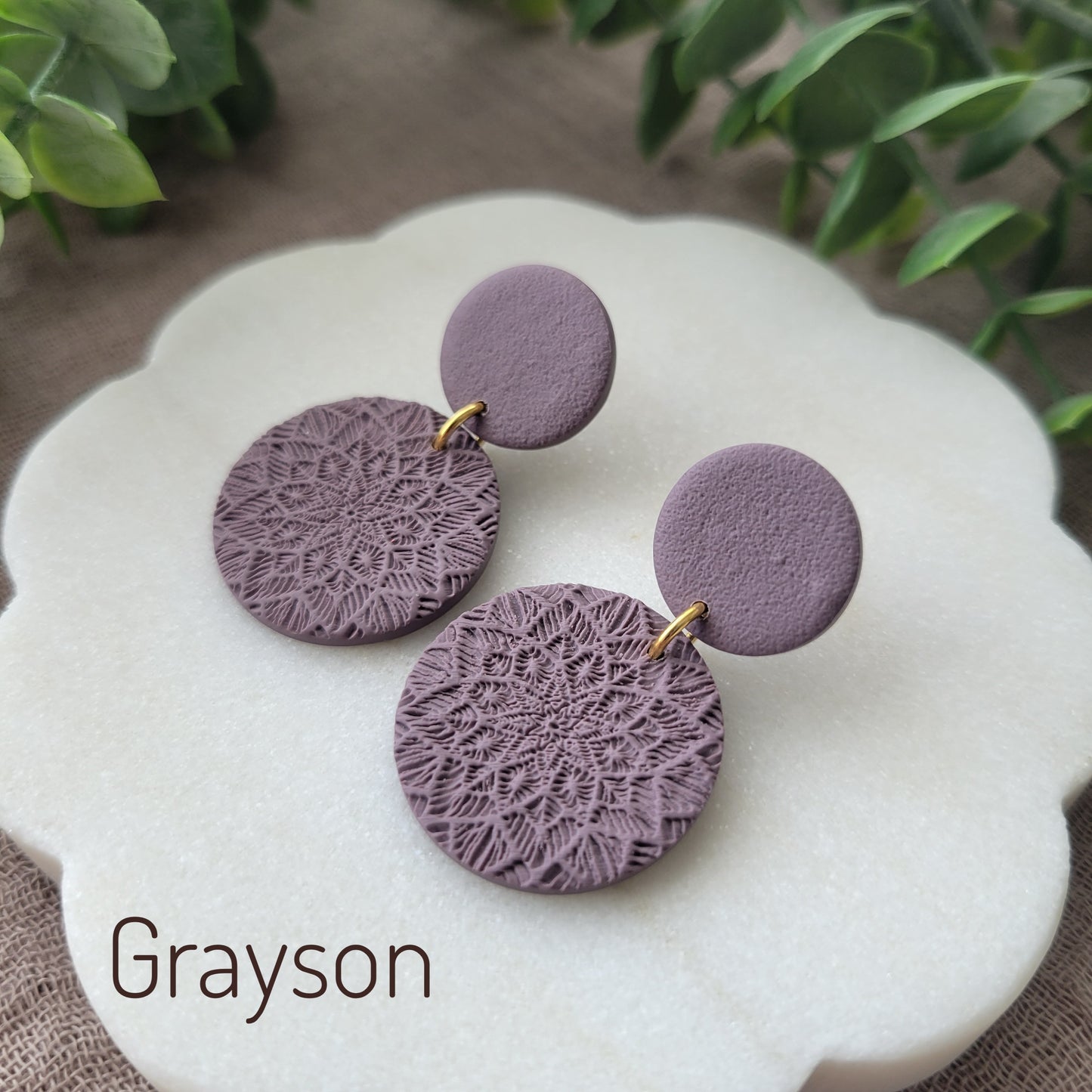 Side view of a pair of purple mandala textured earrings displayed on a white round scalloped tray.