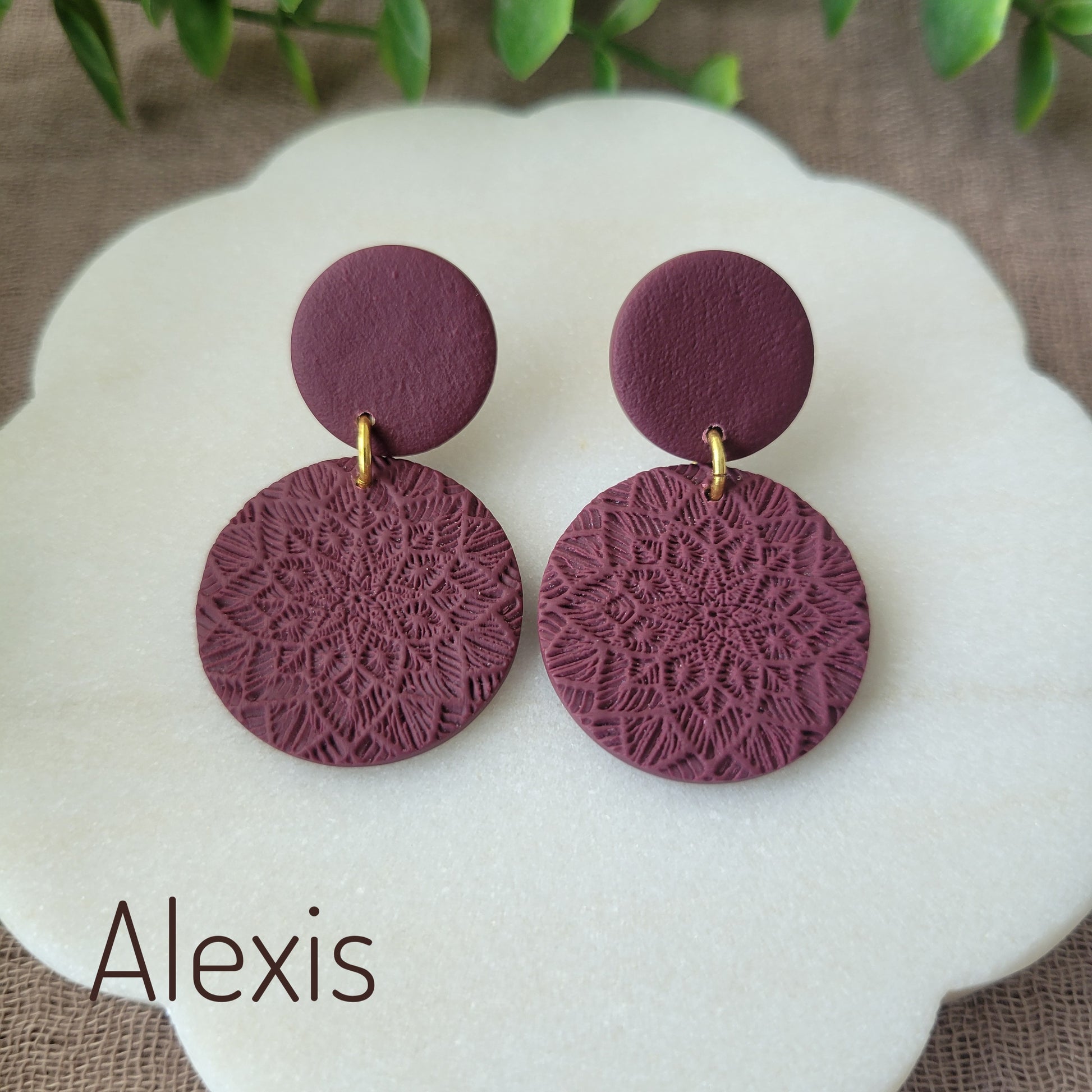 Top view of a pair of reddish purple mandala textured earrings displayed on a white round scalloped tray.