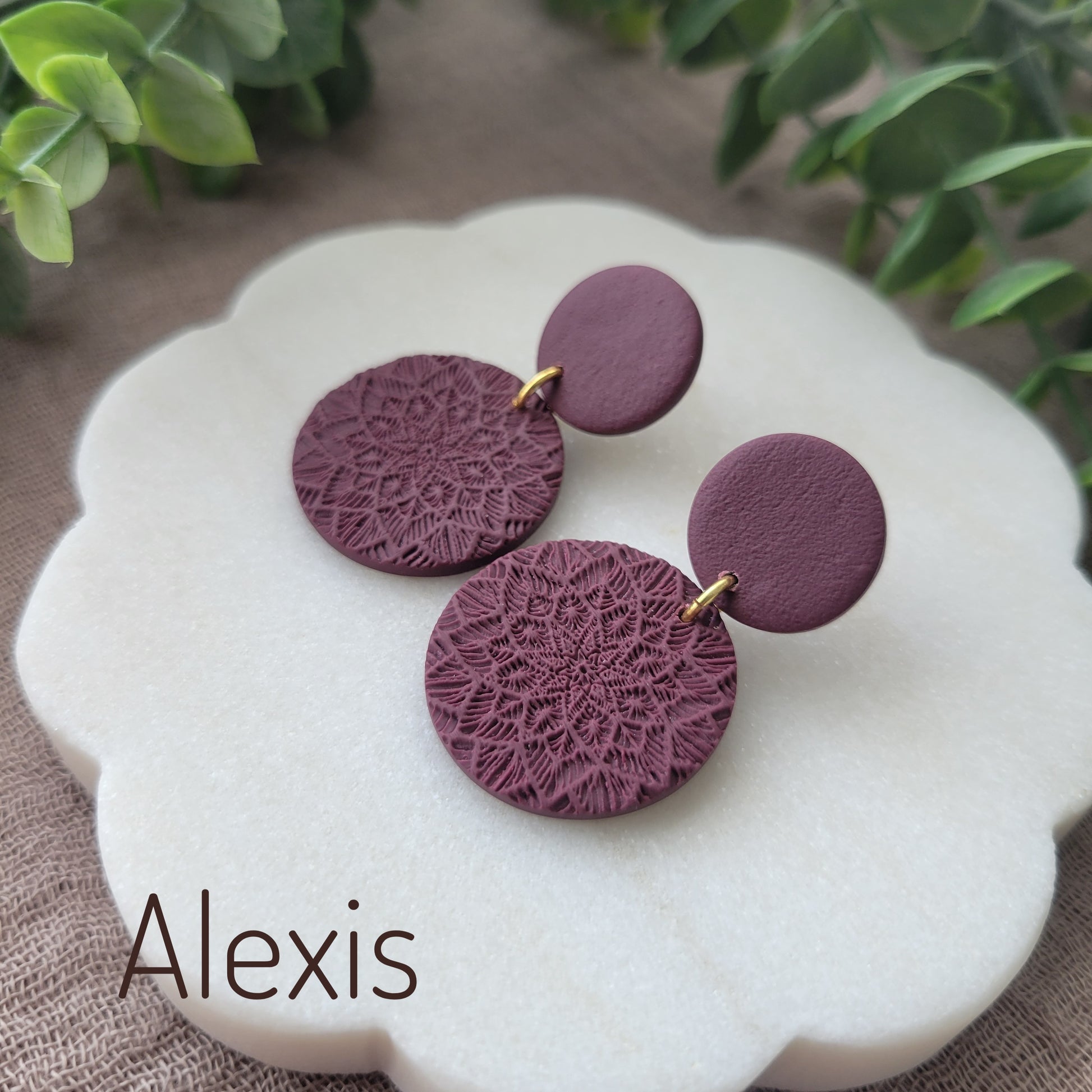 Side view of a pair of reddish purple mandala textured earrings displayed on a white round scalloped tray.