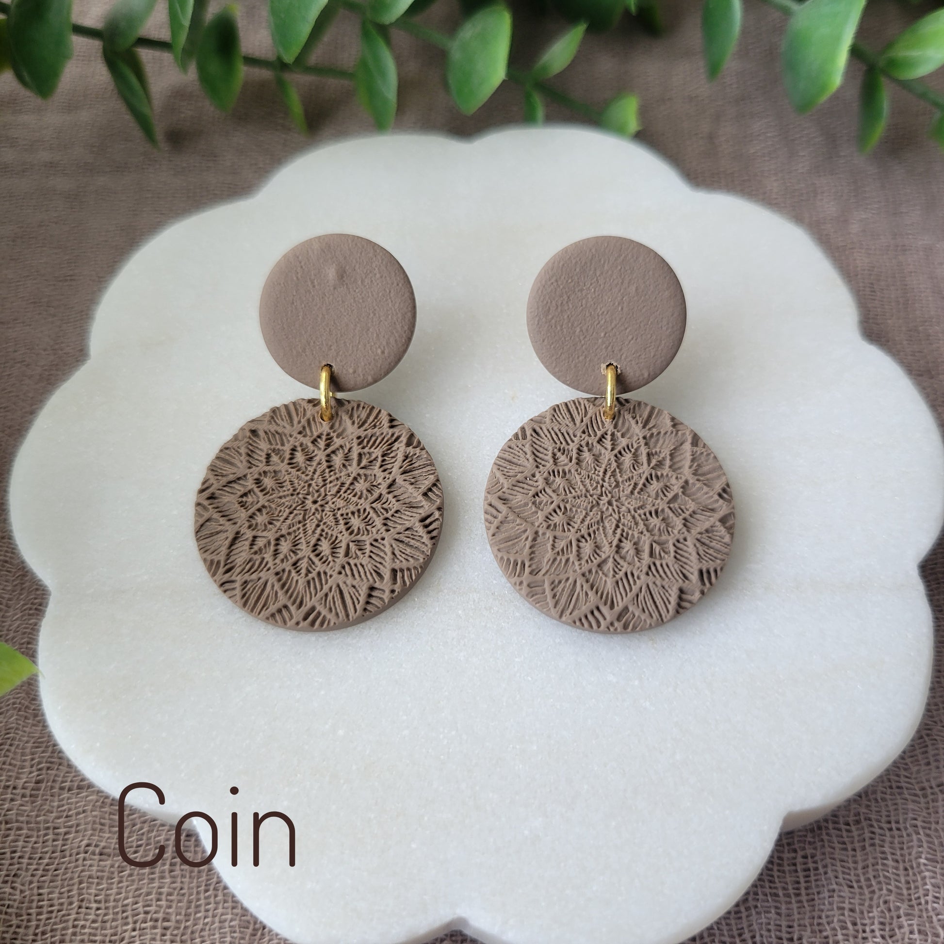 Top view of a pair of brown mandala textured earrings displayed on a white round scalloped tray.