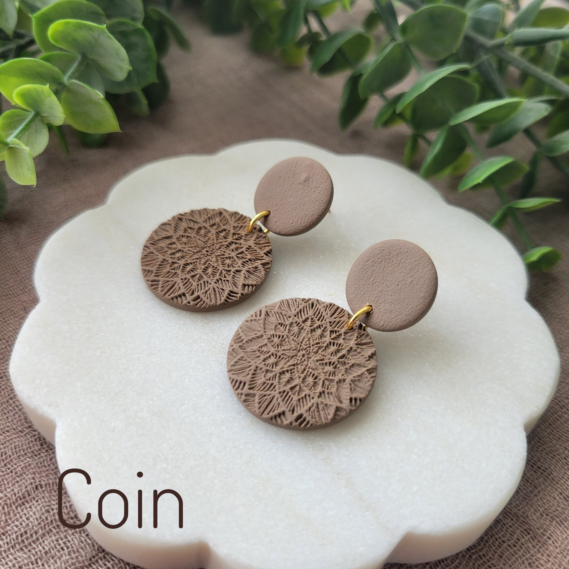 Side view of a pair of brown mandala textured earrings displayed on a white round scalloped tray.