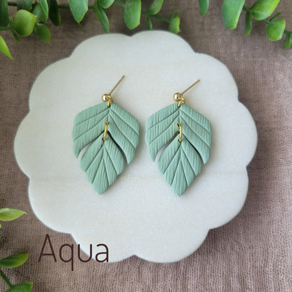 Top view of a pair of greenish blue leaf shaped earrings displayed on a white scalloped tray.