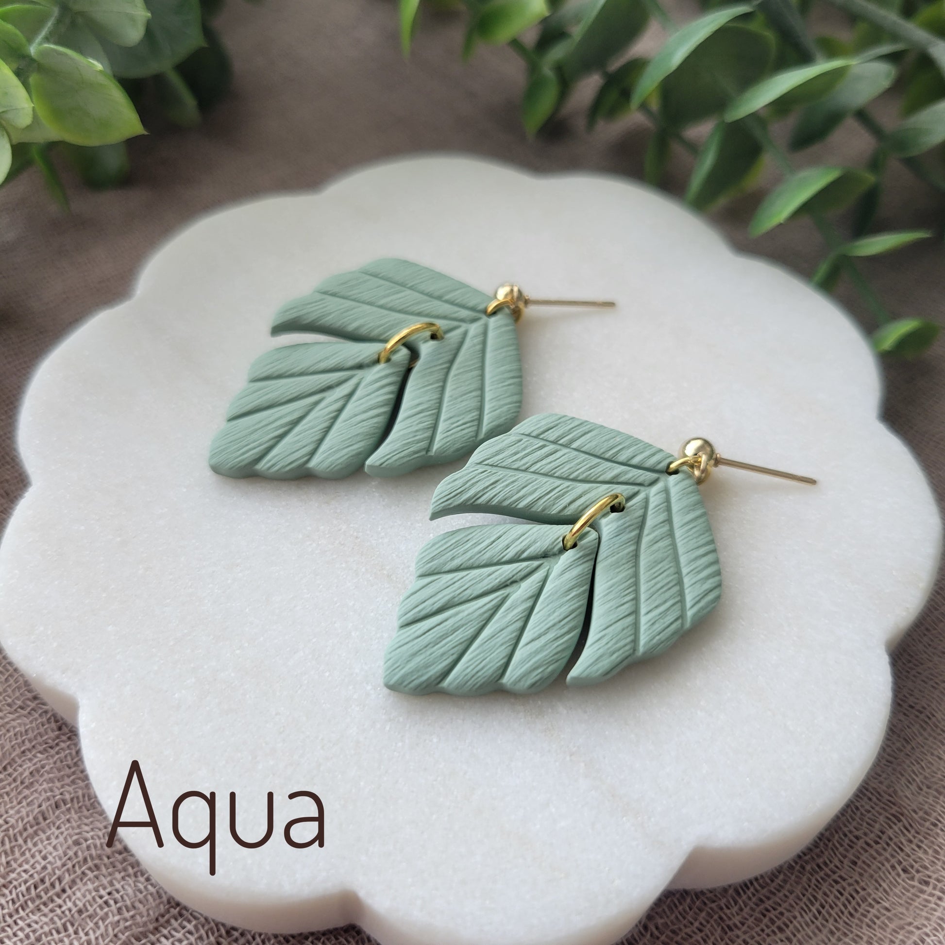 Side view of a pair of greenish blue leaf shaped earrings displayed on a white scalloped tray.