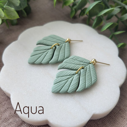 Side view of a pair of greenish blue leaf shaped earrings displayed on a white scalloped tray.