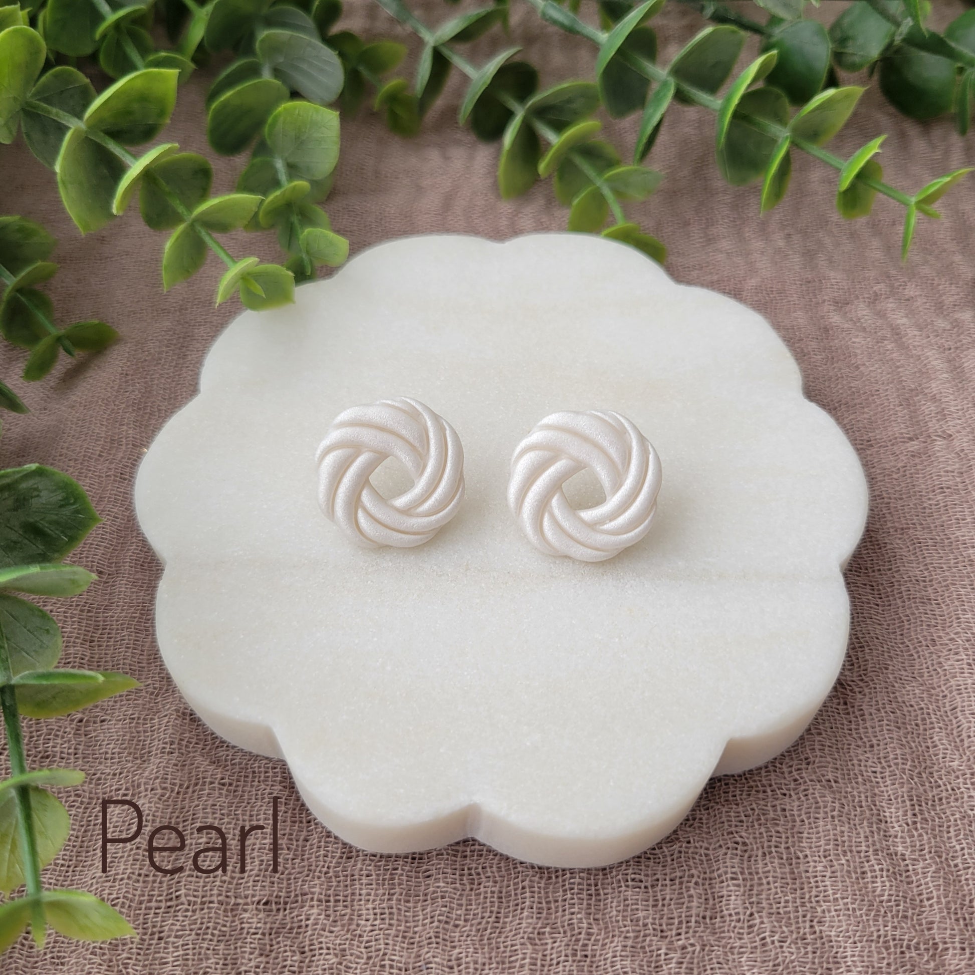 Top view of a pair of pearl knot stud earrings displayed on a white round scalloped tray.