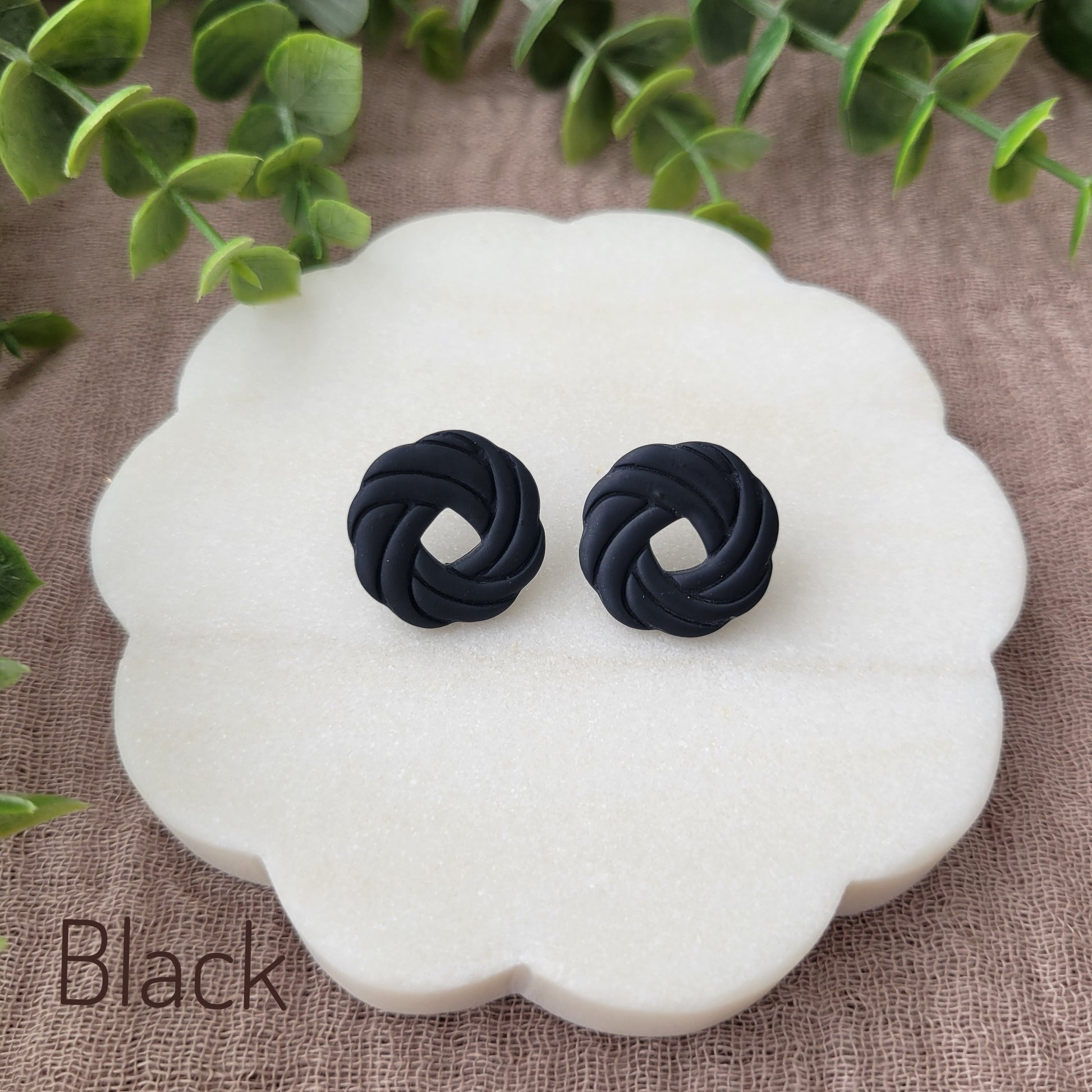 Top view of a pair of black knot stud earrings displayed on a white round scalloped tray.