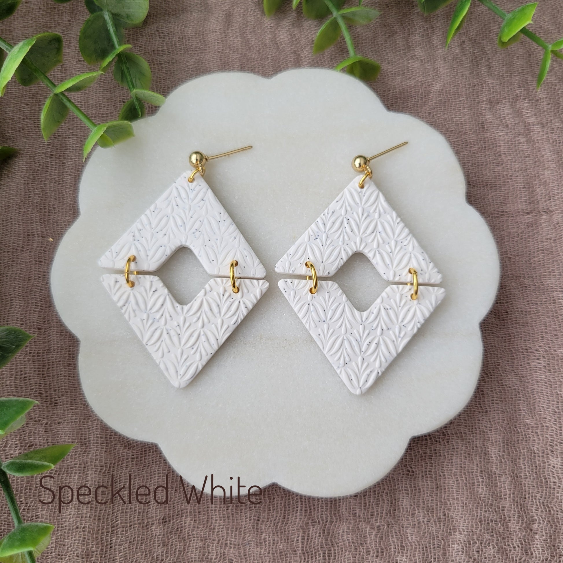 Top view of a pair of speckled white geometric diamond shaped earrings with art deco flower texture displayed on a white scalloped tray.