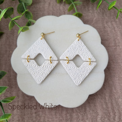 Top view of a pair of speckled white geometric diamond shaped earrings with art deco flower texture displayed on a white scalloped tray.
