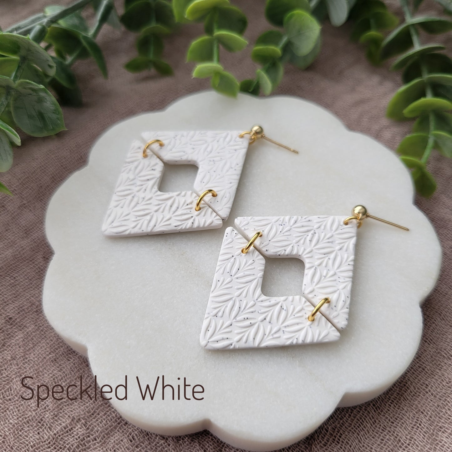Side view of a pair of speckled white geometric diamond shaped earrings with art deco flower texture displayed on a white scalloped tray.