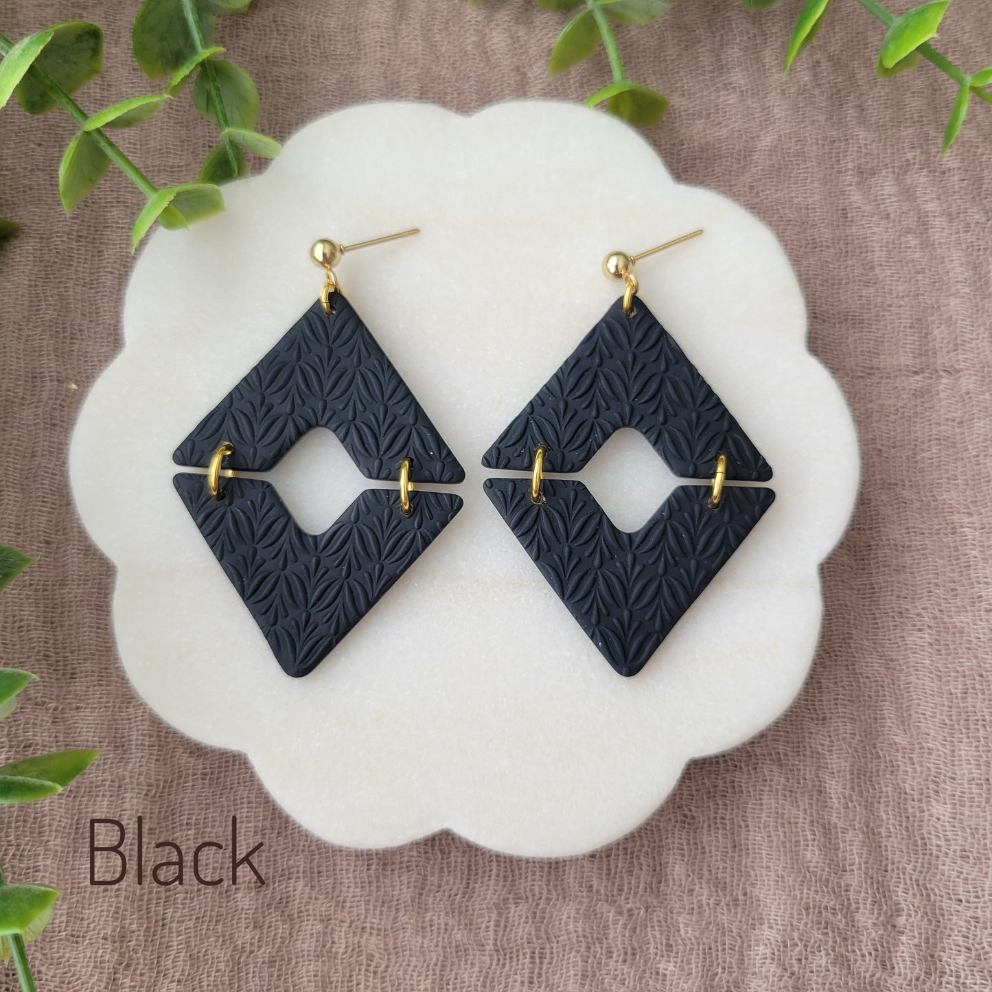 Top view of a pair of black geometric diamond shaped earrings with art deco flower texture displayed on a white scalloped tray.