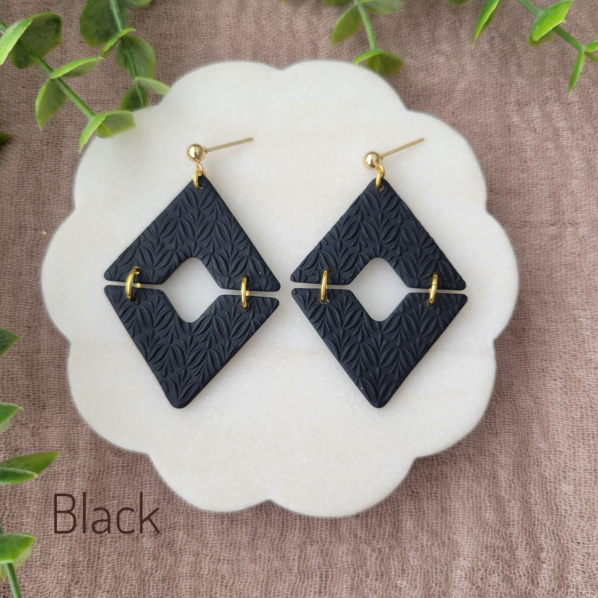 Top view of a pair of black geometric diamond shaped earrings with art deco flower texture displayed on a white scalloped tray.