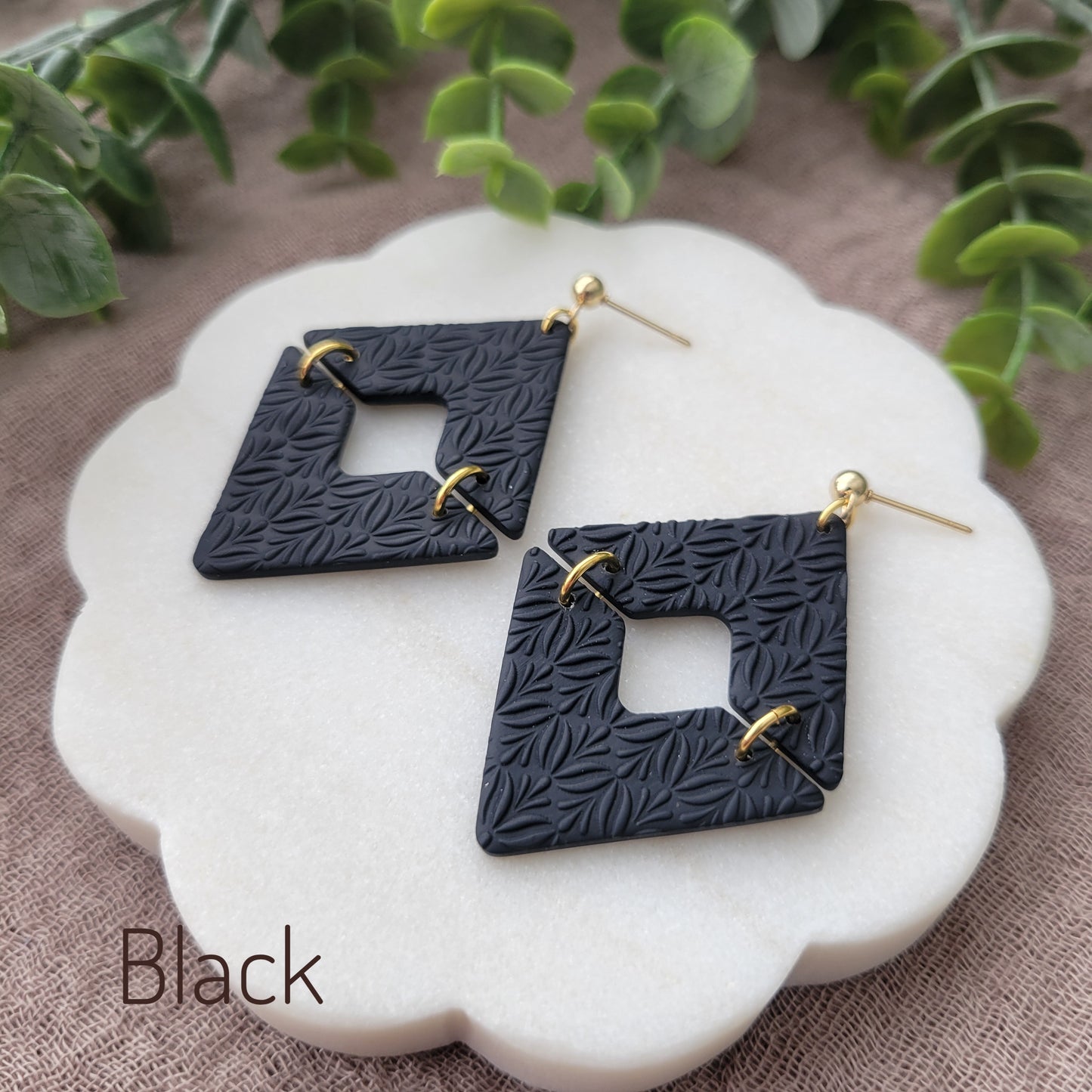 Side view of a pair of black geometric diamond shaped earrings with art deco flower texture displayed on a white scalloped tray.