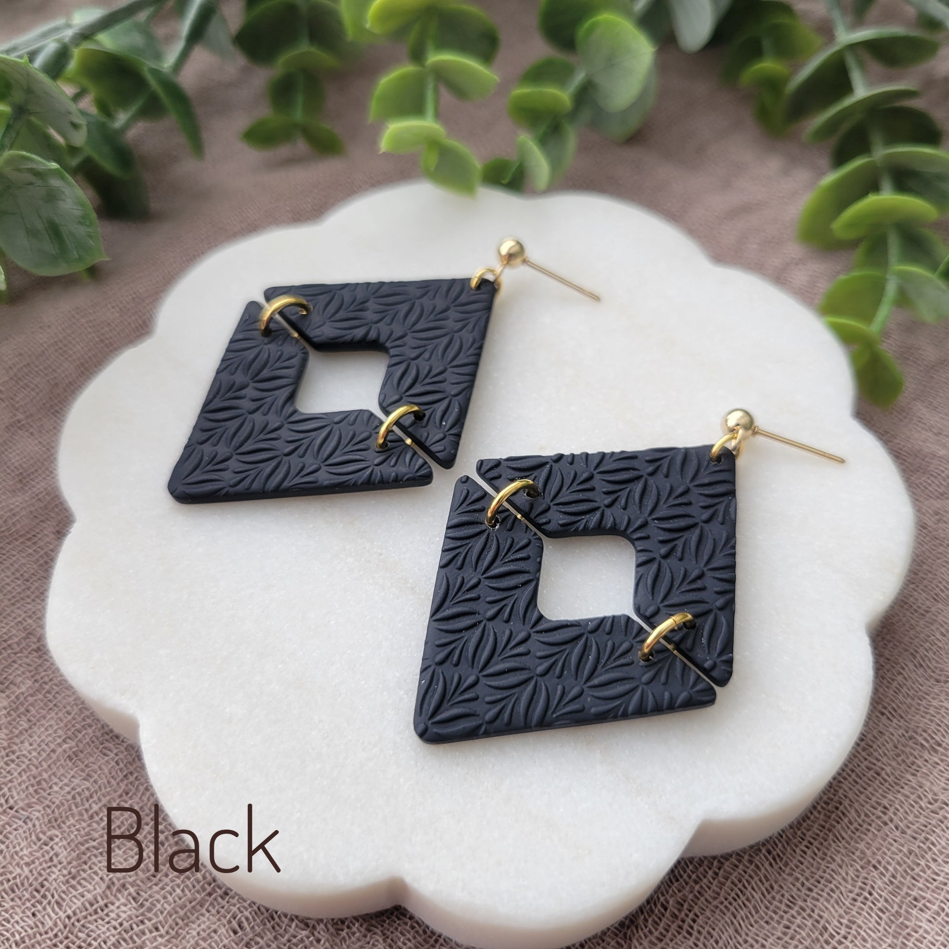 Side view of a pair of black geometric diamond shaped earrings with art deco flower texture displayed on a white scalloped tray.