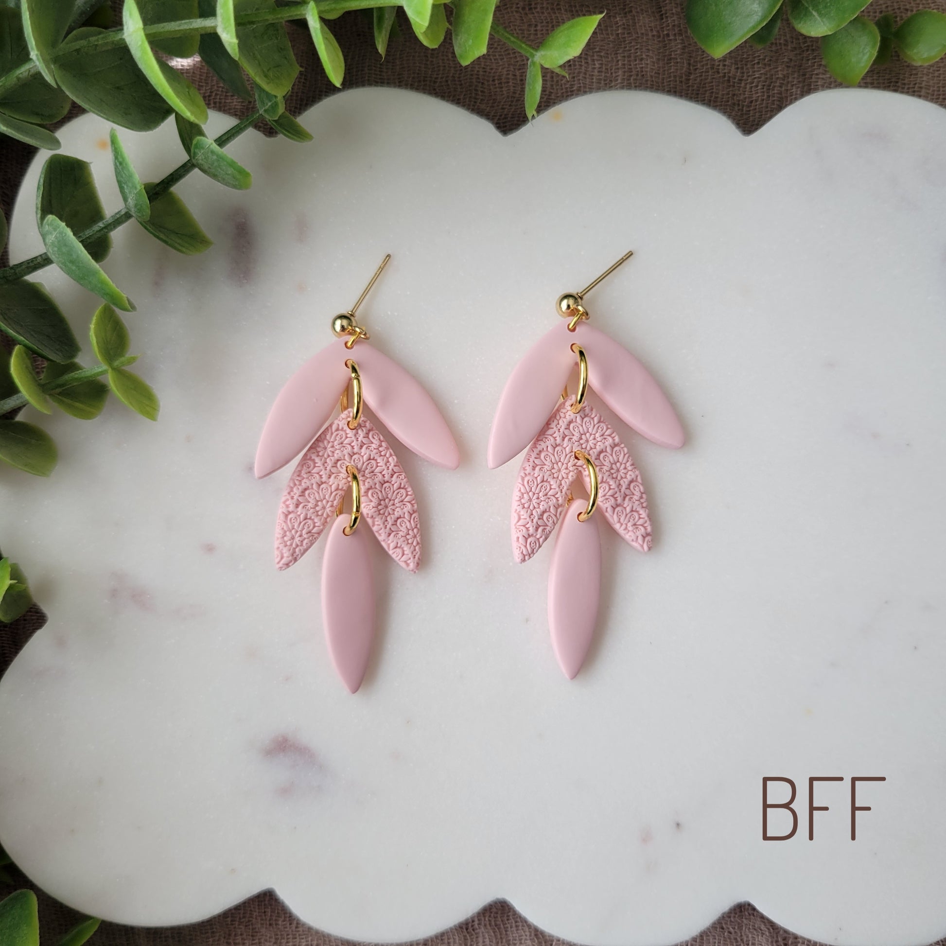 Top view of a pair of pink leaf drop shaped earrings with a scalloped mandala texture displayed on a white scalloped tray.