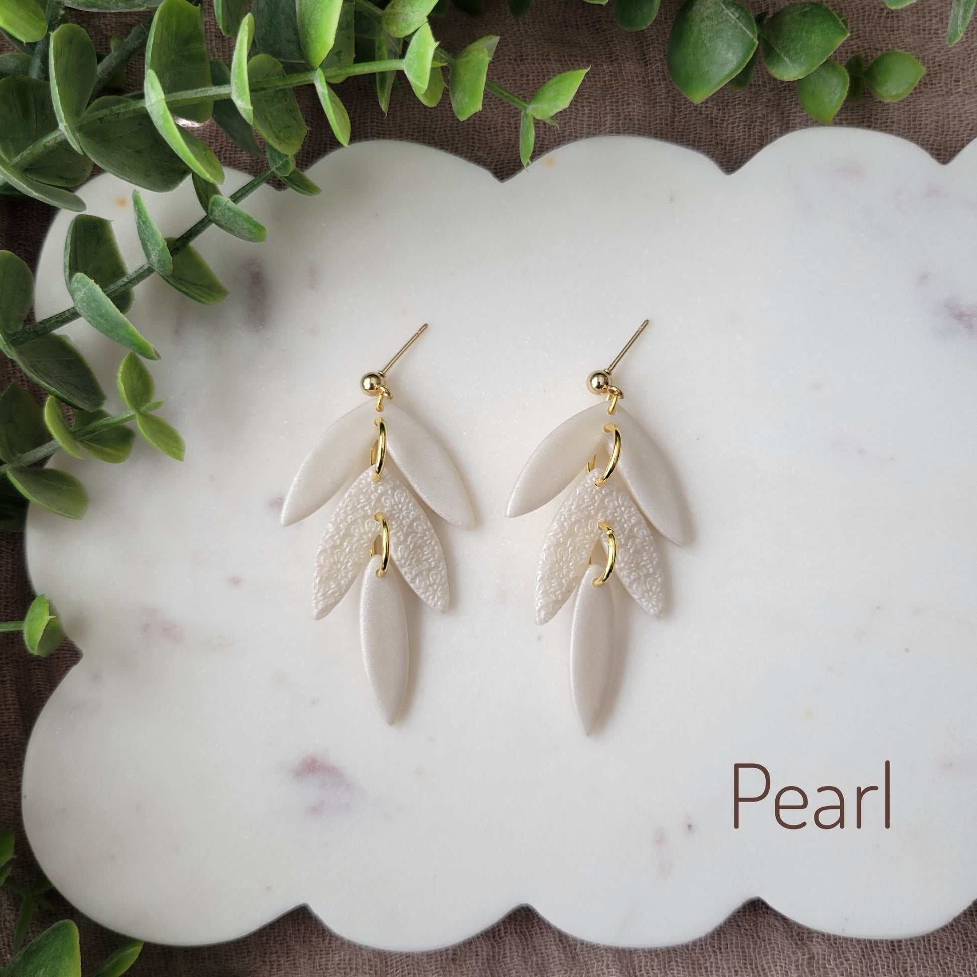 Top view of a pair of pearl leaf drop shaped earrings with a scalloped mandala texture displayed on a white scalloped tray.
