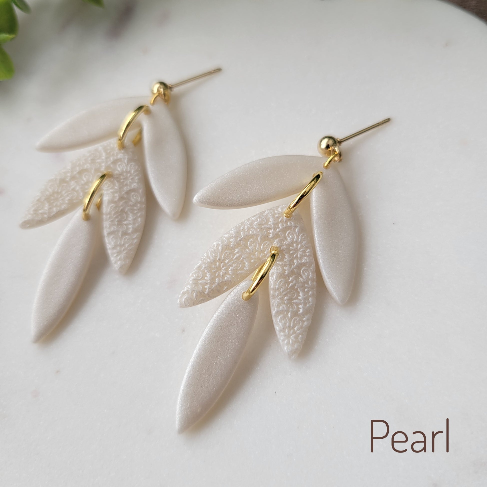 Close up side view of a pair of pearl leaf drop shaped earrings with a scalloped mandala texture displayed on a white scalloped tray.