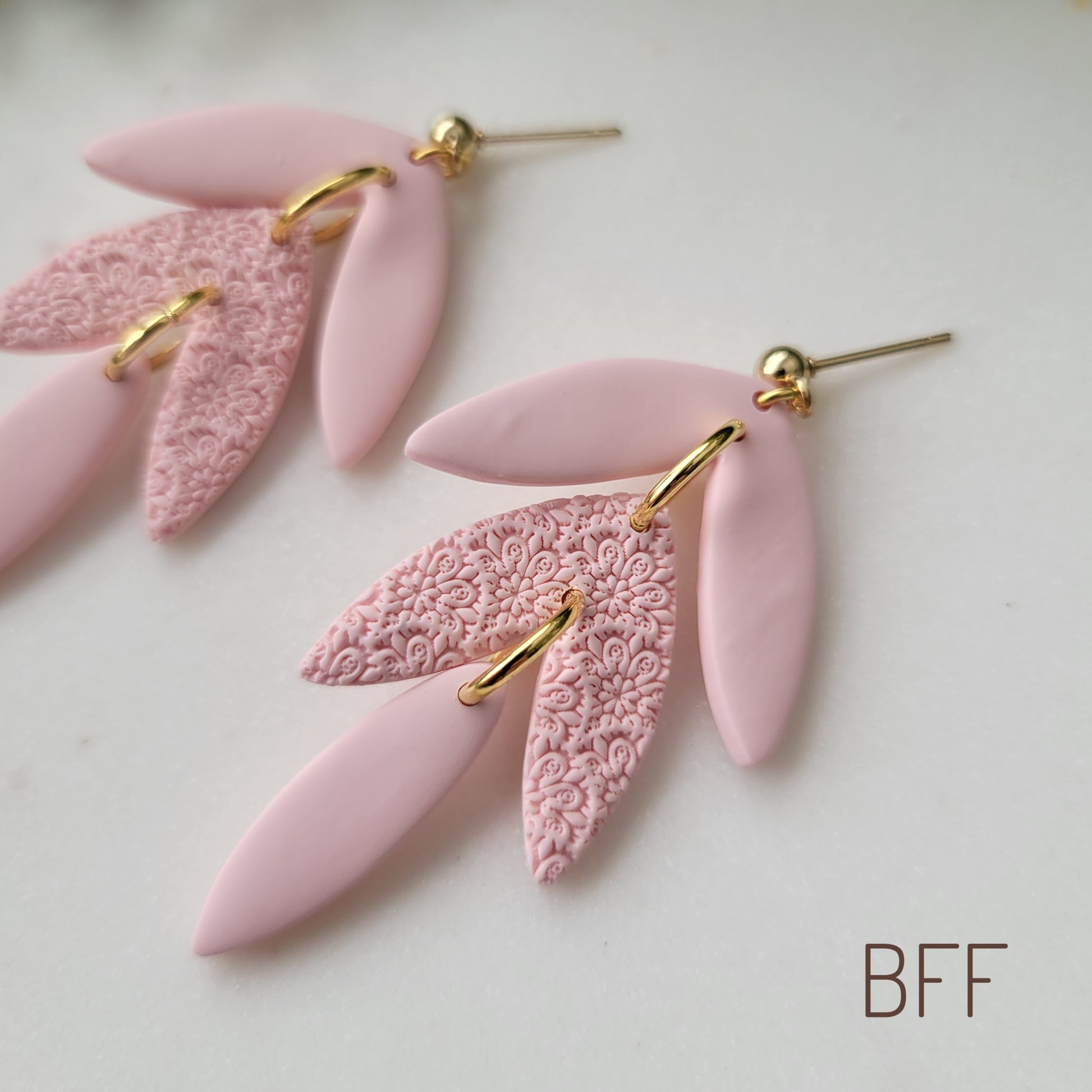 Close up side view of a pair of pink leaf drop shaped earrings with a scalloped mandala texture displayed on a white scalloped tray.