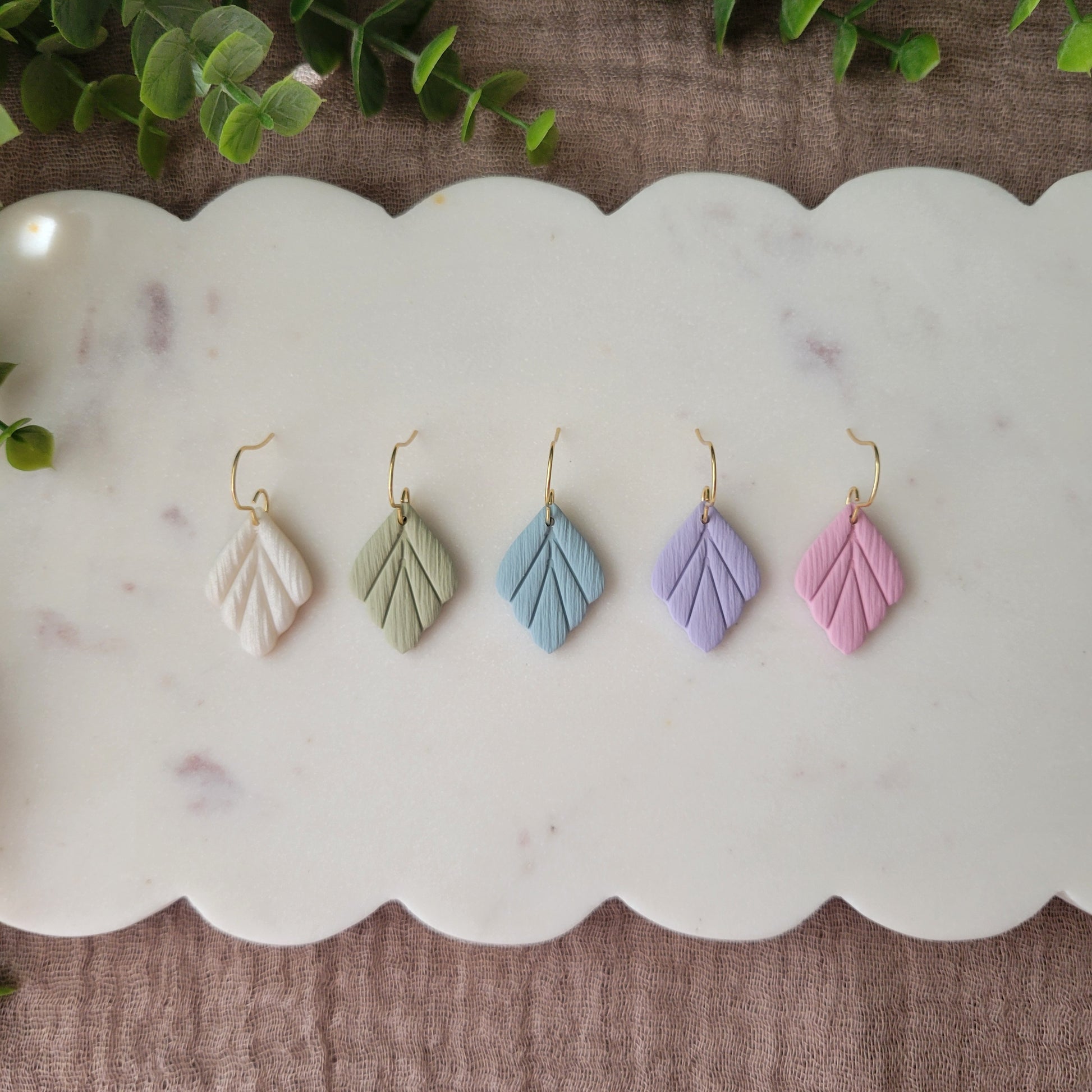 Top view of five small leaf shaped earrings displayed on a white scalloped tray.