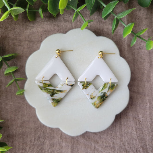 Top view of a pair of white, green, gold accented geometric diamond earrings displayed on a white round scalloped tray.
