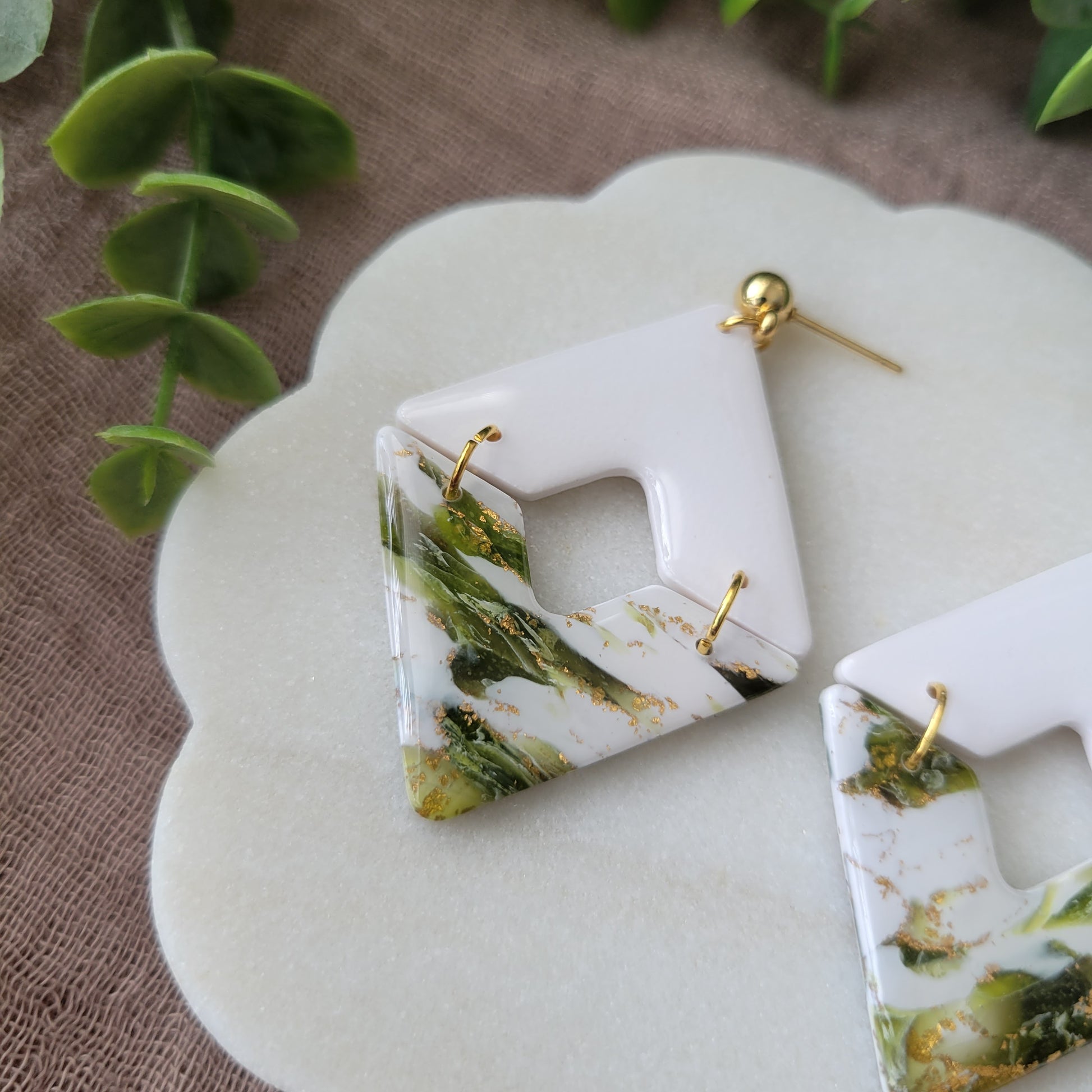 Close up view of a pair of white, green, gold accented geometric diamond earrings displayed on a white round scalloped tray.