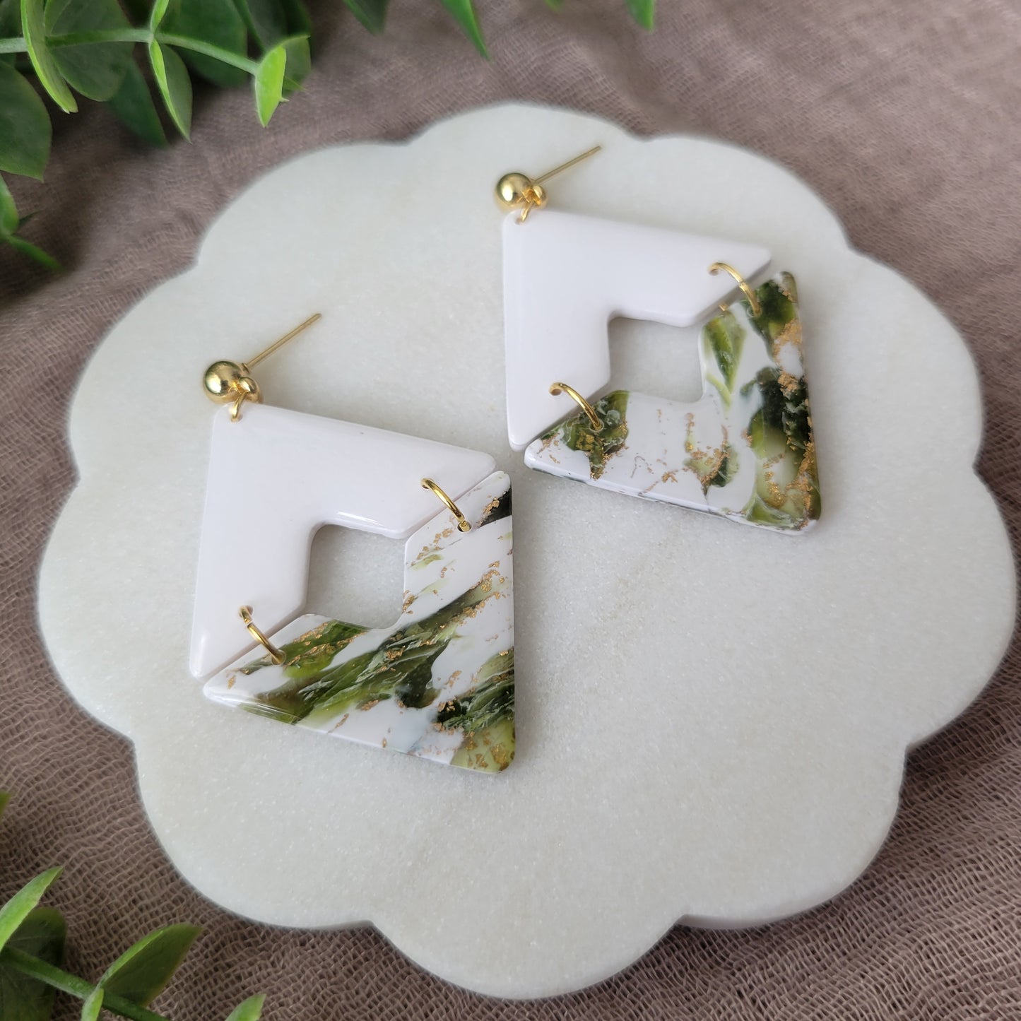 Left side view of a pair of white, green, gold accented geometric diamond earrings displayed on a white round scalloped tray.