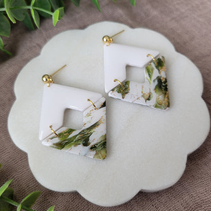 Left side view of a pair of white, green, gold accented geometric diamond earrings displayed on a white round scalloped tray.