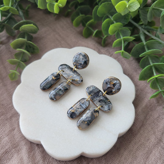 Right side view of a pair of gray, gold stone shaped earrings displayed on a white round scalloped tray.