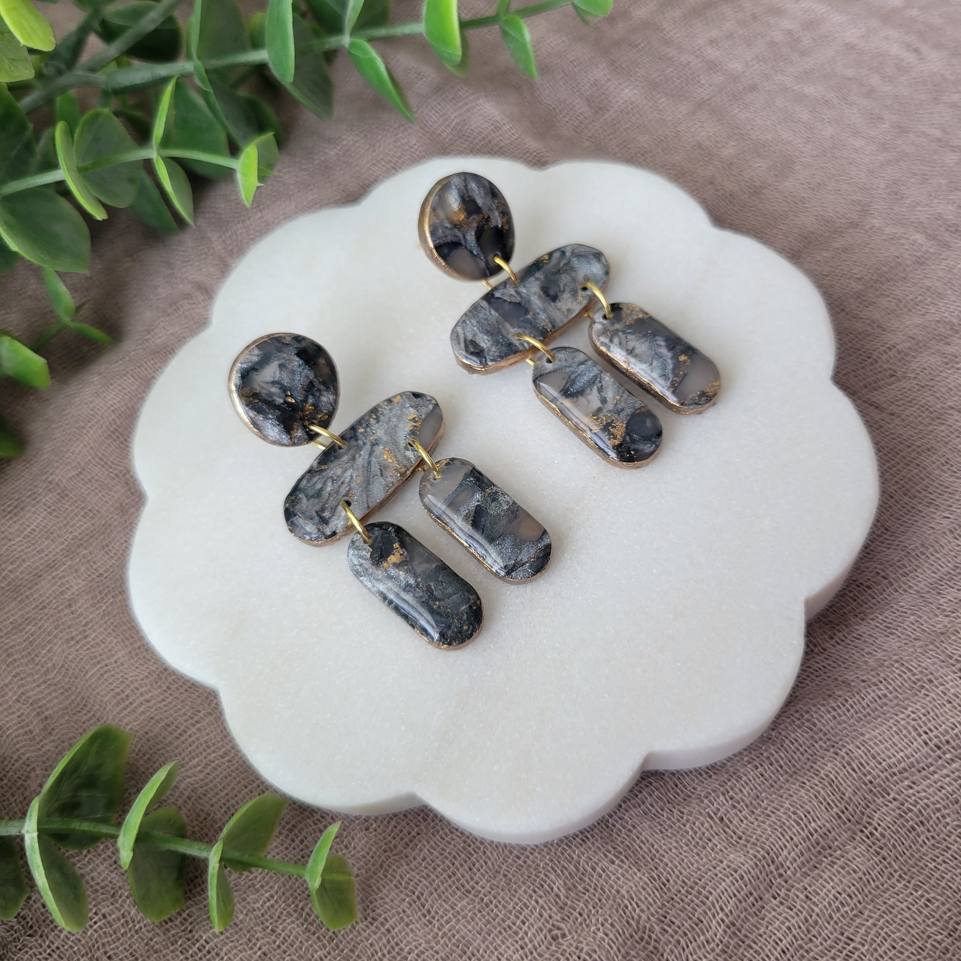 Left side view of a pair of gray, gold stone shaped earrings displayed on a white round scalloped tray.