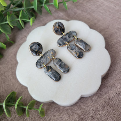 Left side view of a pair of gray, gold stone shaped earrings displayed on a white round scalloped tray.