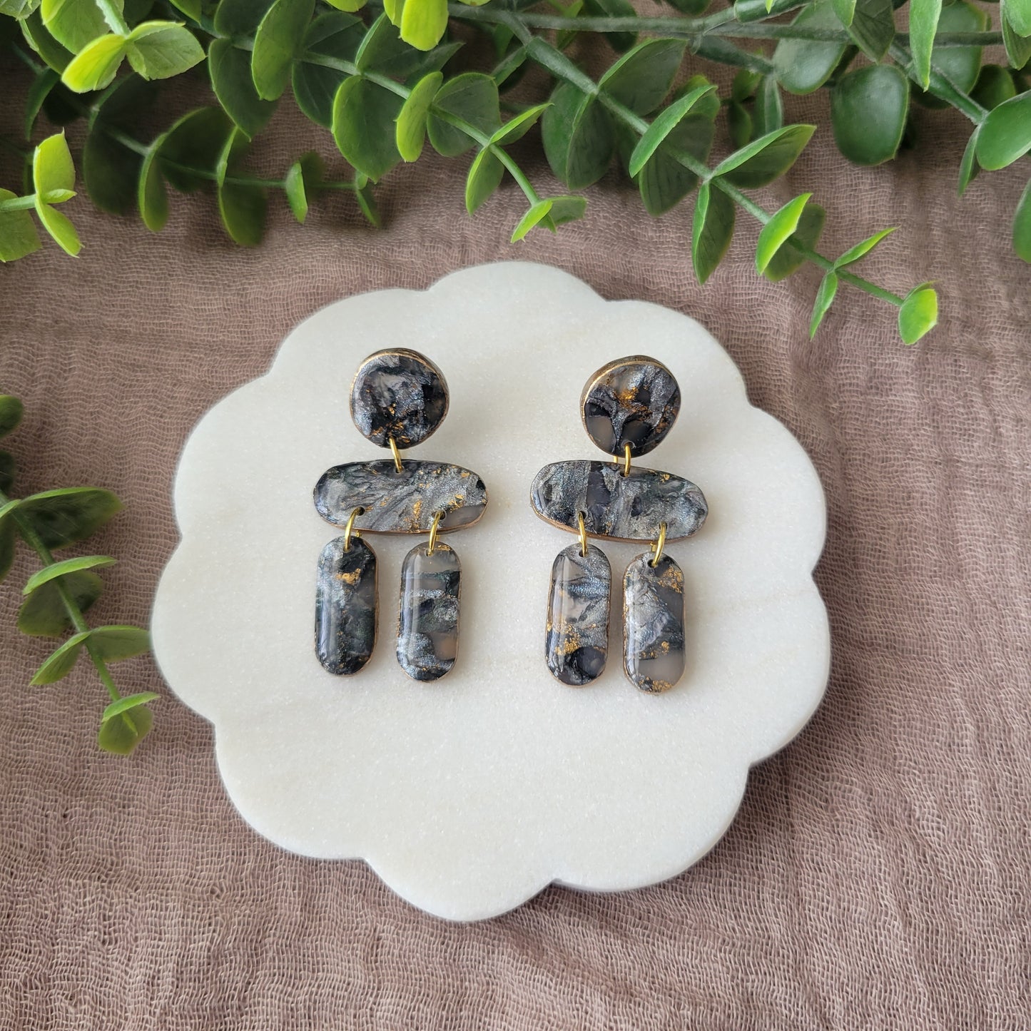 Top view of a pair of gray, gold stone shaped earrings displayed on a white round scalloped tray.