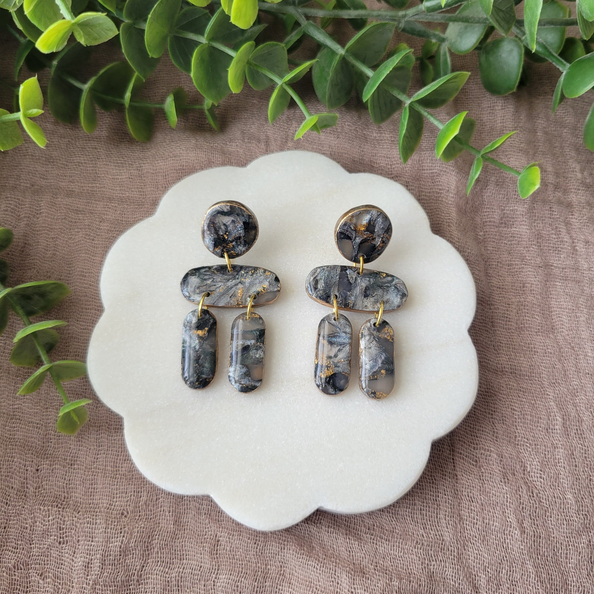 Top view of a pair of gray, gold stone shaped earrings displayed on a white round scalloped tray.