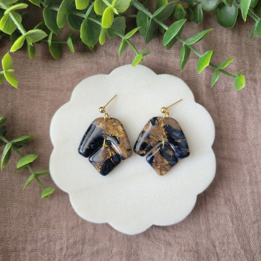 Top view of a pair of dark blue, bronze marble boomerang shaped earrings displayed on a white round scalloped tray.