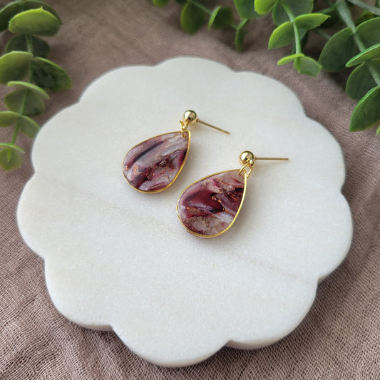 Right side view of a pair of maroon, pearl teardrop earrings with a gold trim bezel displayed on a white round scalloped tray.