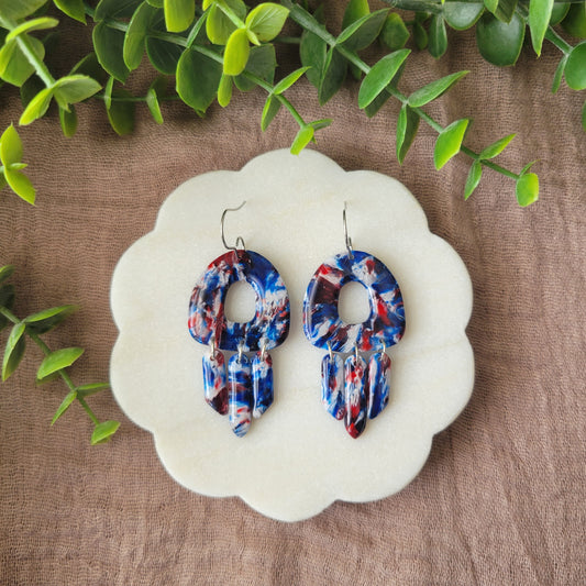 Top view of a pair of red, white, blue marble earrings displayed on a white round scalloped tray.