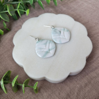 Left side view of a pair of  tiffany blue, white marble embossed round tear drop shaped earrings displayed on a white round scalloped tray.