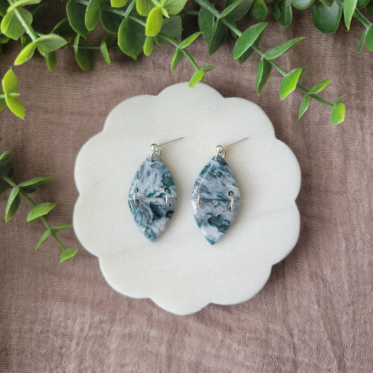 Top view of a pair of green, white, silver marble earrings displayed on a white round scalloped tray.