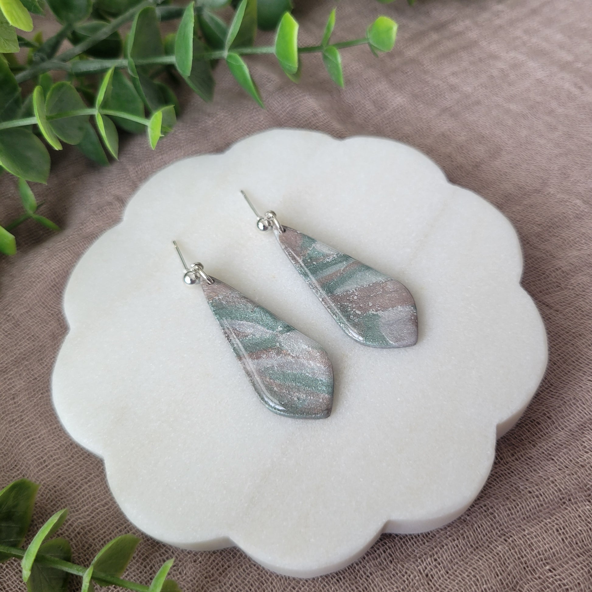 Left side view of a pair of shimmering pastel elongated tear drop shaped earrings displayed on a white round scalloped tray.
