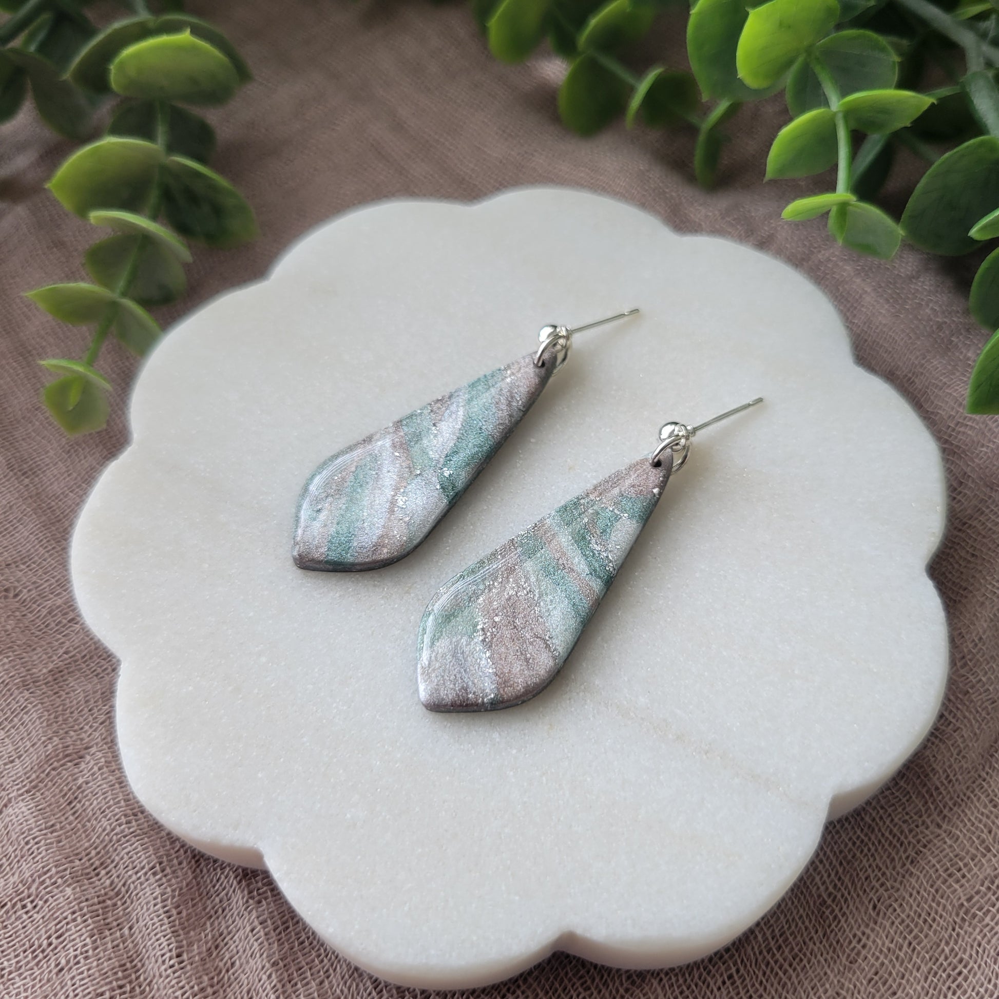 Right side view of a pair of shimmering pastel elongated tear drop shaped earrings displayed on a white round scalloped tray.