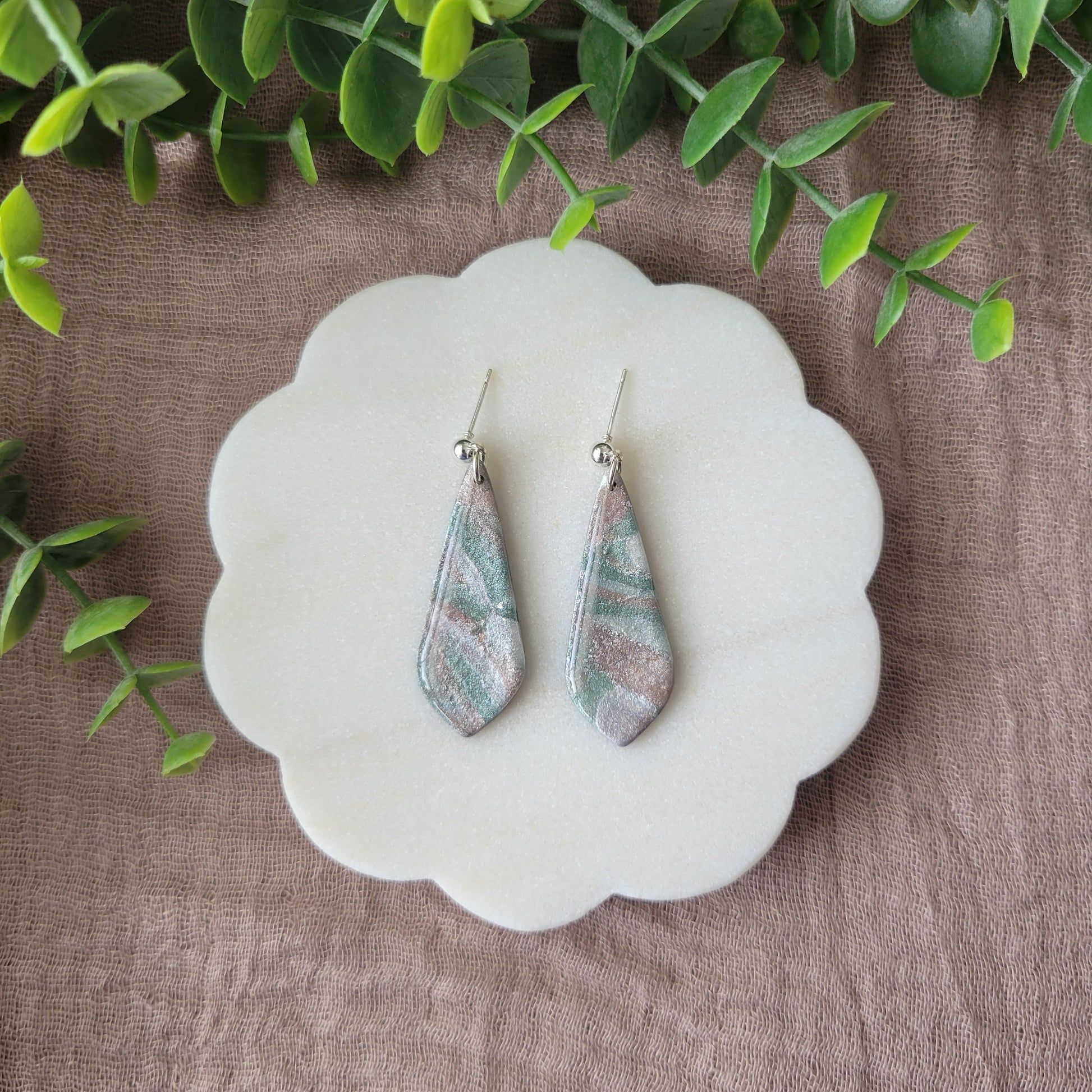 Top view of a pair of shimmering pastel elongated tear drop shaped earrings displayed on a white round scalloped tray.