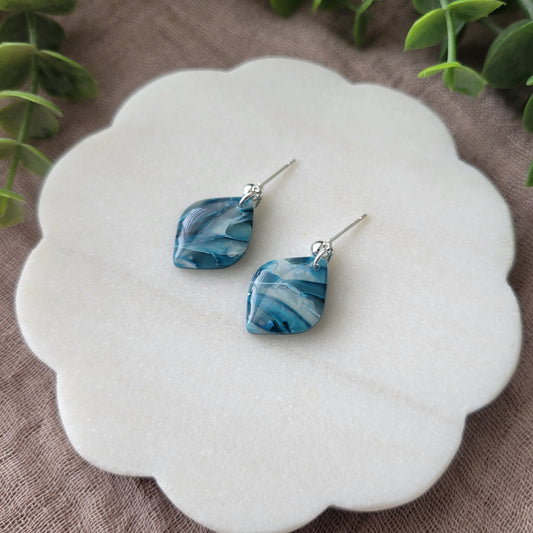 Right side view of a pair of blue marble diamond shaped earrings displayed on a white round scalloped tray.