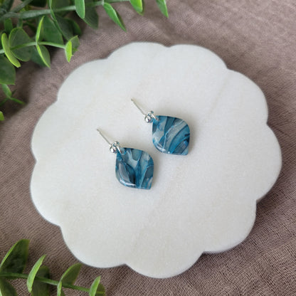 Left side view of a pair of blue marble diamond shaped earrings displayed on a white round scalloped tray.