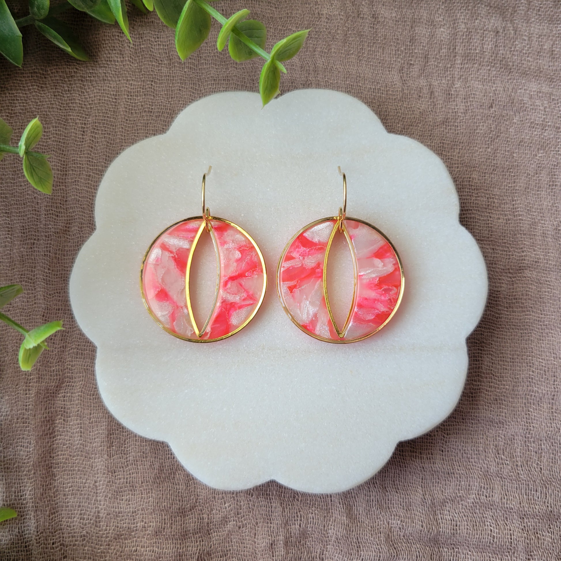 Top view of a pair of round polymer clay earrings in the color bright pink and pearl with gold trim on a white scalloped tray.