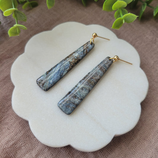 Right side view of a pair of gray, brown pillar shaped earrings displayed on a white round scalloped tray.