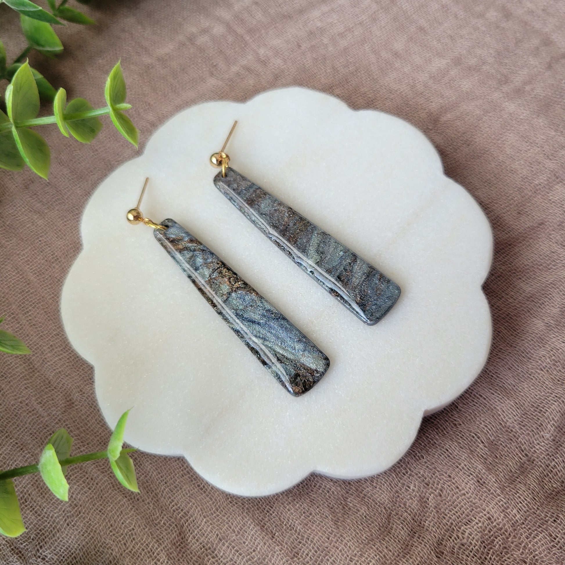 Left side view of a pair of gray, brown pillar shaped earrings displayed on a white round scalloped tray.