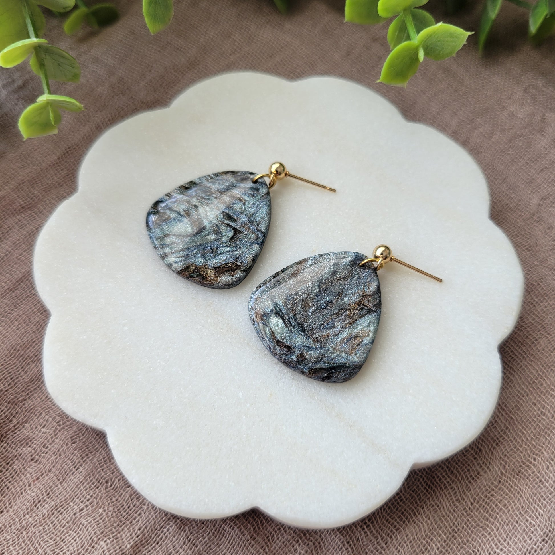 Right side view of a pair of gray, brown guitar pick shaped earrings displayed on a white round scalloped tray.