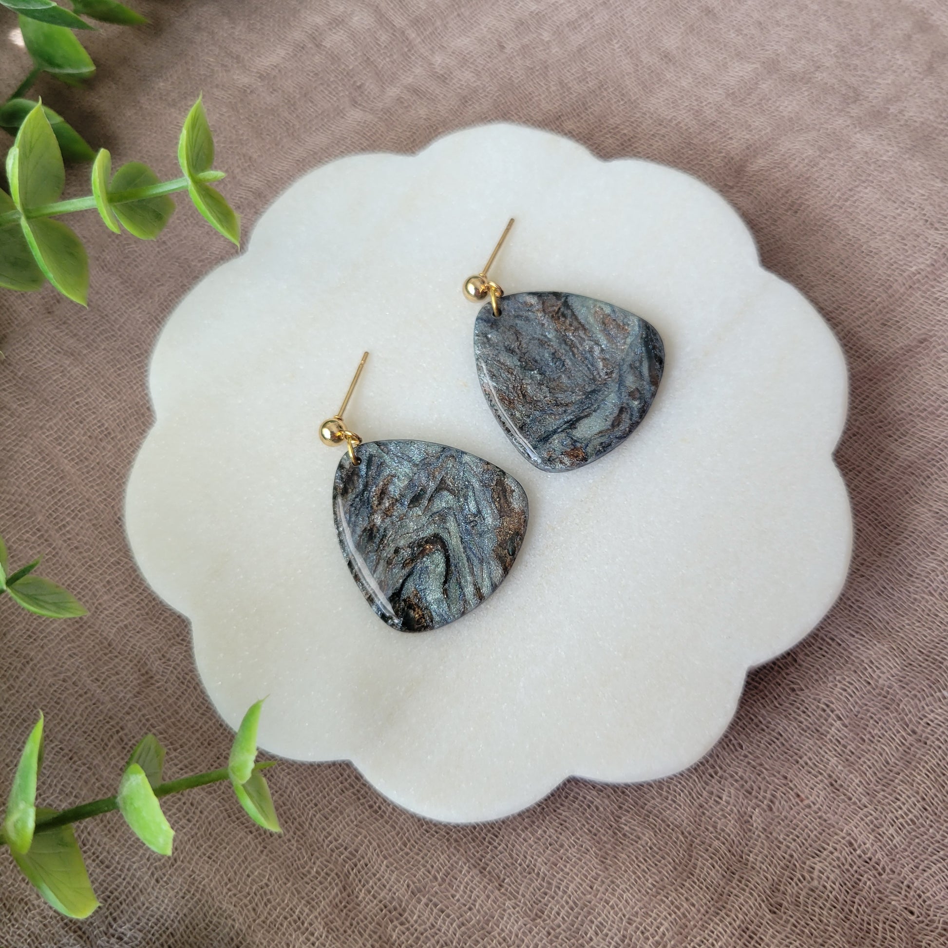 Left side view of a pair of gray, brown guitar pick shaped earrings displayed on a white round scalloped tray.